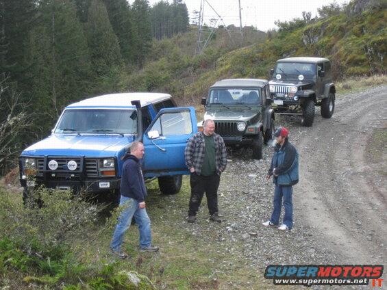 Blue Ford Bronco with snorkel and white roof parked on gravel road with two Jeeps behind.