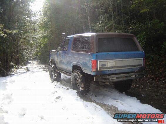 Blue Ford Bronco on a snowy trail in the woods, rear quarter view.