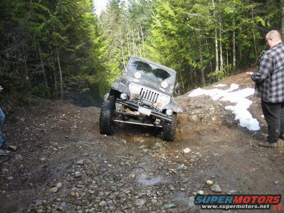 Jeep Wrangler tilted on a muddy forest trail with two people watching.