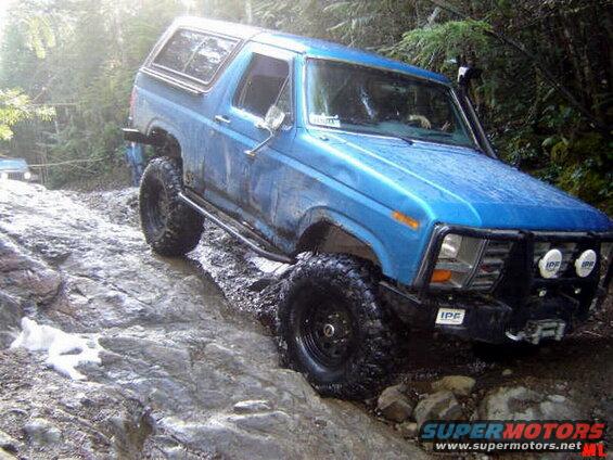 Blue 1980s Ford Bronco off-roading on a rocky, wet trail.