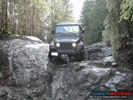 Dark green Jeep Wrangler climbing a steep rocky and muddy trail in a forest.