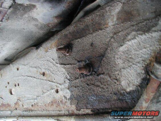 Close-up of two rusty bolts on the underside of a vehicle frame with textured undercoating.