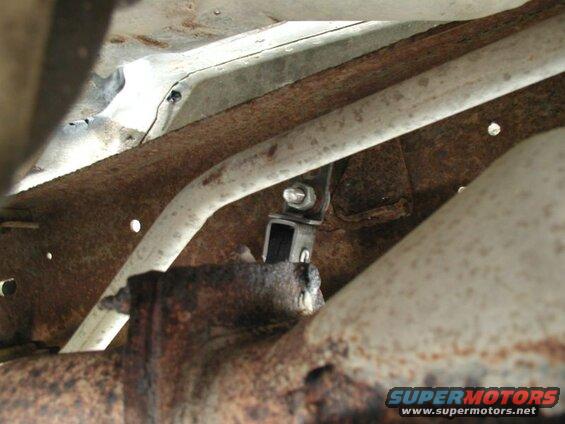 Underneath a rusted vehicle frame, a metal exhaust pipe is visible with a new black bracket attached.