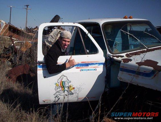 Guy sticking tongue out, giving thumbs up in doorway of old white pickup truck in junkyard
