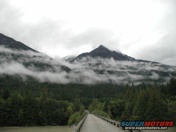 Bridge leading to a forest and misty mountains.