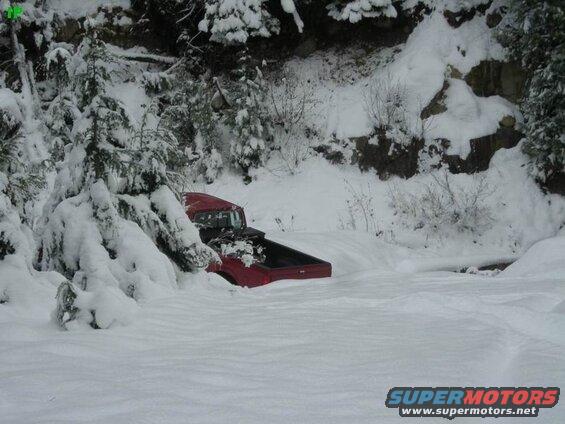 Red Ford pickup truck stuck in deep snow on a wooded mountainside.