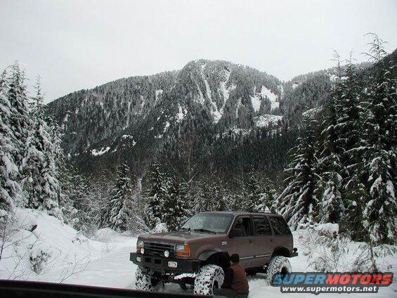 Brown Ford Explorer SUV with large tires in a snowy forest.