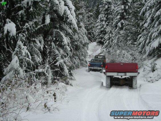 Red Ford F-250 driving on a snow-covered road in a forest, with two other trucks behind it.