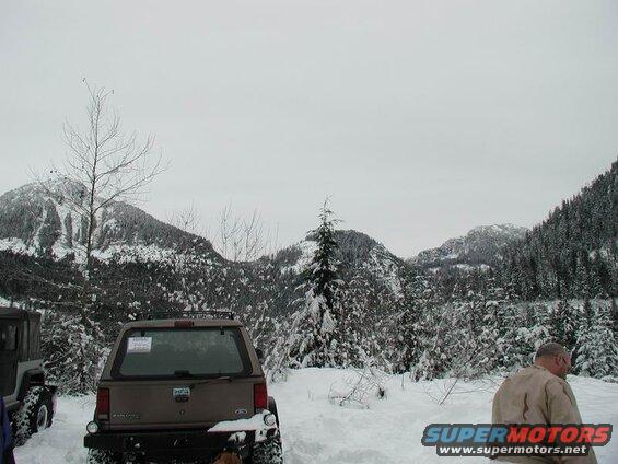 Brown Ford SUV parked in snow with mountains and trees behind it.