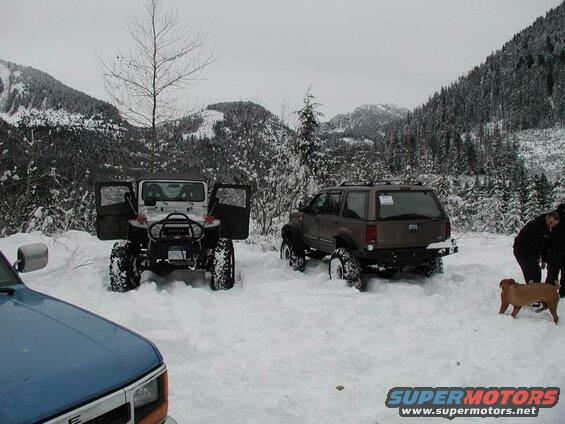 Two off-road vehicles in deep snow with mountains and trees in the background. A dog and two people are also visible.