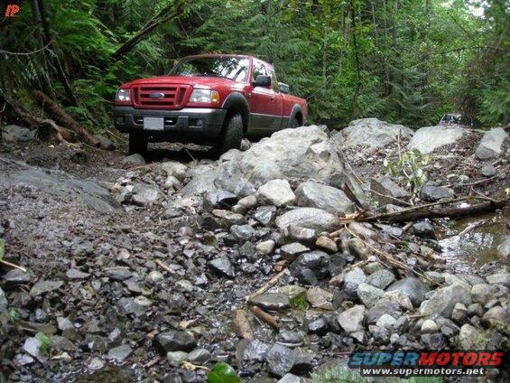 Red Ford pickup truck driving on a rocky, wooded trail.