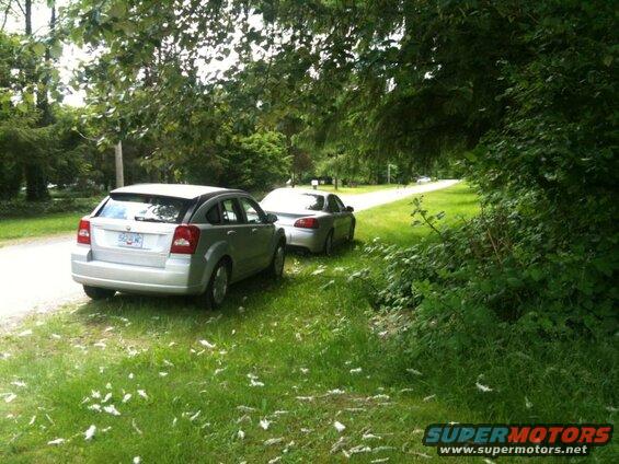 Silver hatchback parked on a gravel road next to a grassy area with trees.