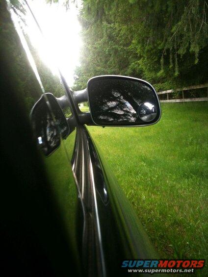 Black car side mirror reflecting trees and sky, parked on grass next to trees and fence.