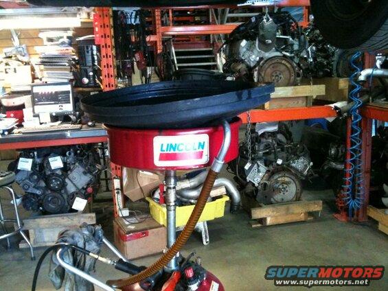 Red Lincoln oil drain pan with black top in a garage with car engines in the background.