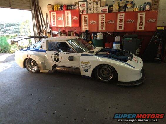 White and blue custom racing car with large rear wing and exposed engine in a garage.