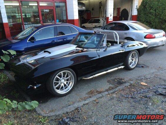 Black convertible Corvette with silver hood scoop and side pipes parked next to other cars.