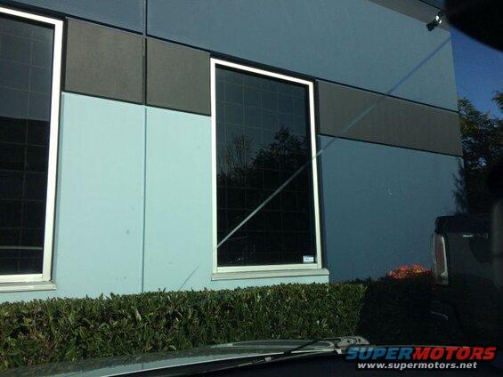 Exterior of a blue building with dark windows and a hedge. Part of a dark vehicle is visible.