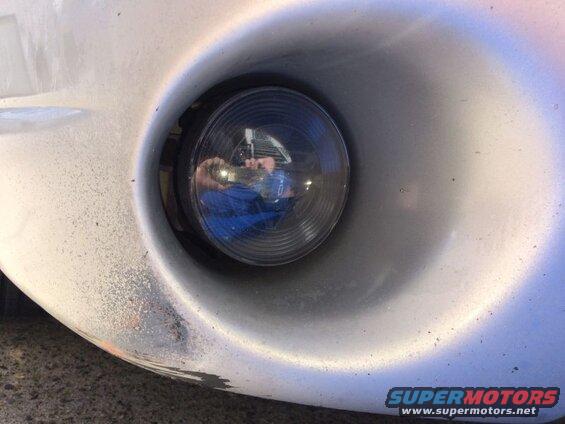 Close-up of a silver car's fog light housing with a dark fog light lens reflecting a person.