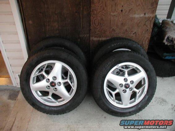 Four silver alloy wheels with tires stacked in front of a wooden wall.