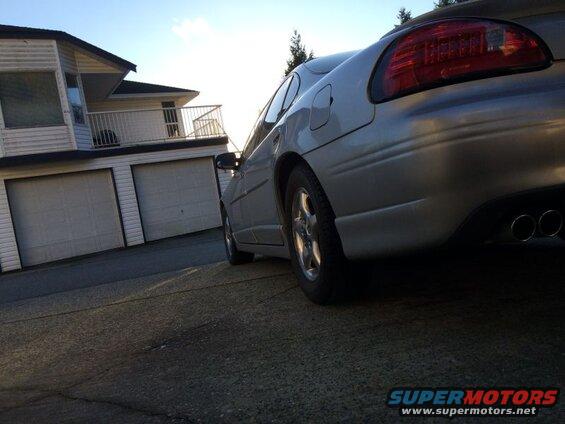 Silver sedan parked in front of a house, showing rear driver's side and dual exhaust.
