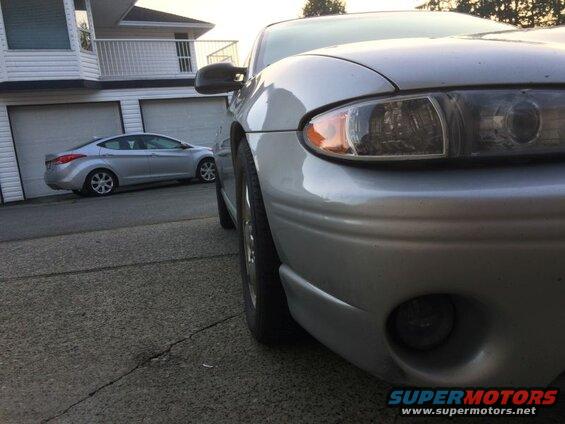 Close-up of silver sedan front fender and headlight, with another sedan in background.