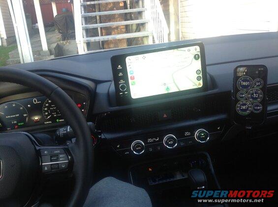 Car interior showing dashboard, touchscreen, smartphone with gauges, and steering wheel.