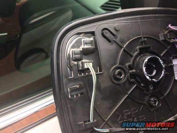 Back of a black side mirror housing with electrical connectors and wires, reflection of side mirror glass visible.