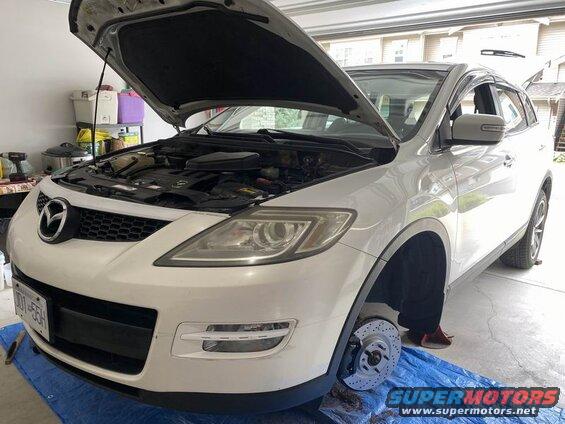 White Mazda CX-9 SUV with hood open and front wheel removed, showing brake components.