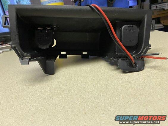 Black plastic automotive dash panel with two power outlets and wires.