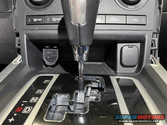 Center console of a vehicle with gear shifter, 12V outlets, and climate control buttons.