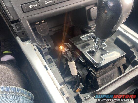 Close-up of car interior center console with disassembled trim and a glowing orange light bulb near the gear shifter.