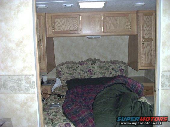 RV bedroom with a floral headboard, plaid comforter, and wooden cabinets above the bed