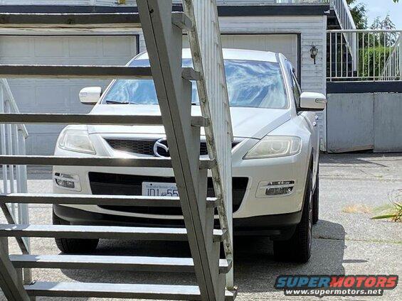 White Mazda CX-9 parked in front of a garage, viewed through metal staircase slats.