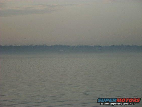 Water with a distant treeline under a hazy sky. SUPERMOTORS.NET logo in lower right