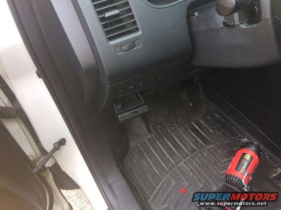 Driver's side interior of a vehicle with dashboard, steering column, floor mat, and a red power tool.