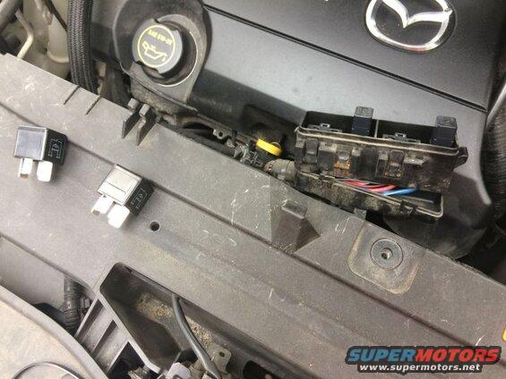 Two automotive relays on a black plastic engine cover next to an open fuse box.