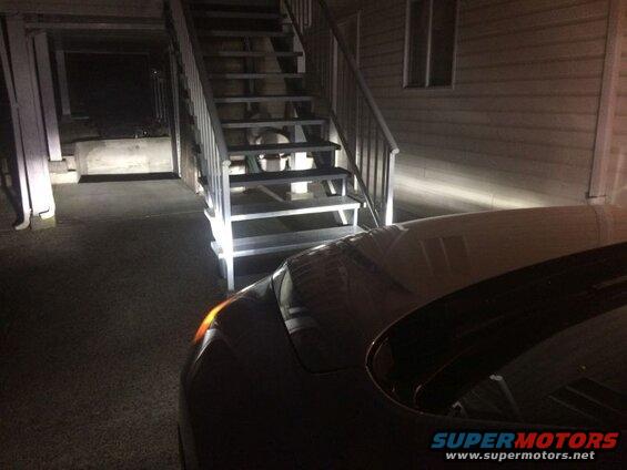 Front corner of a dark car with illuminated turn signal next to outdoor stairs.