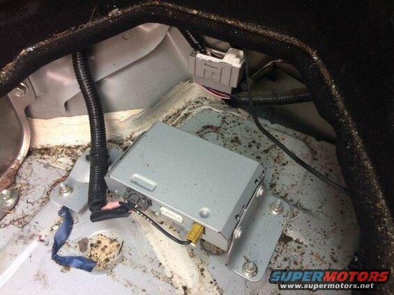 Silver electronic module with wires and conduit in a dirty vehicle trunk.