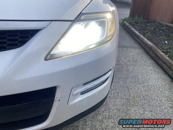 Front right headlight and bumper of a white SUV, headlight is on.