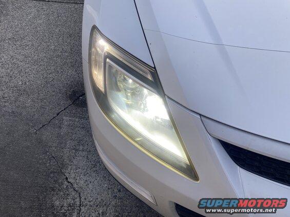 Illuminated headlight of a white SUV parked on cracked asphalt.