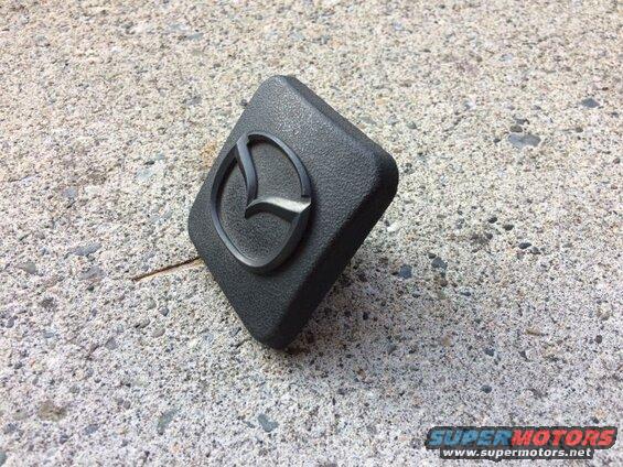 Black Mazda emblem on a dark gray automotive part on concrete.