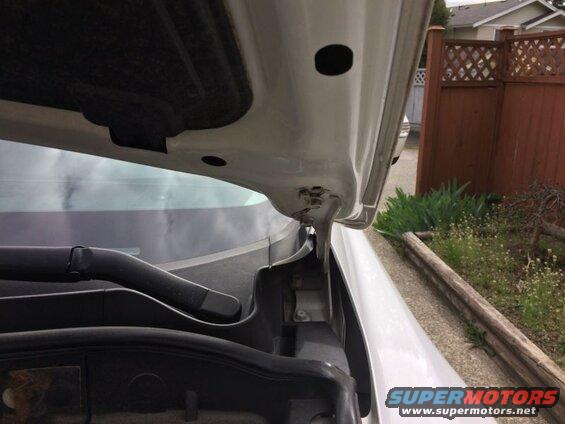 Close-up of a white car hood interior, showing insulation and hinge, with a windshield and outdoor scene beyond.