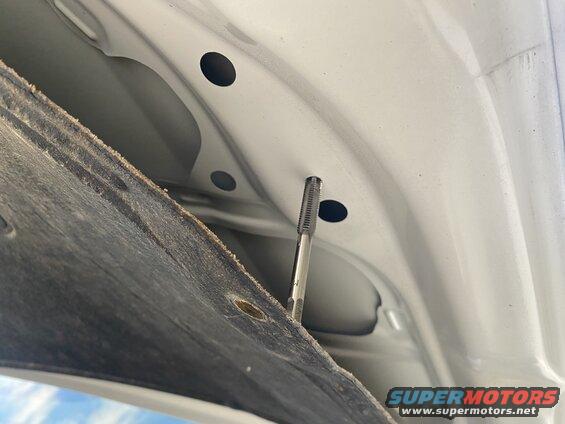 Metal tap tool inserted into a hole on the underside of a white car hood.