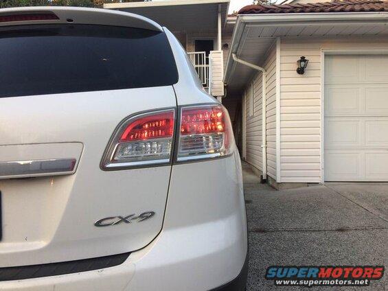 White Mazda CX-9 SUV parked next to a beige garage, rear passenger side view.