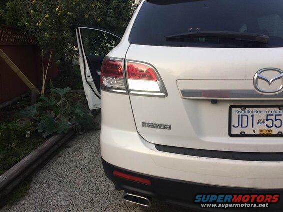 White Mazda CX-9 SUV rear driver's side with rear door open, parked on gravel next to a garden.