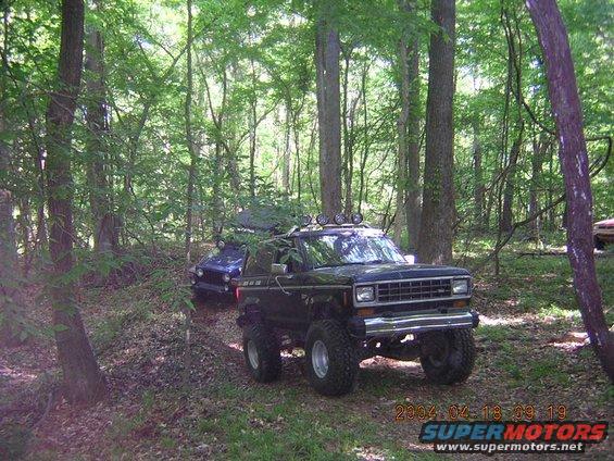 Lifted black Ford Bronco II off-roading in woods with a blue Jeep Cherokee behind it