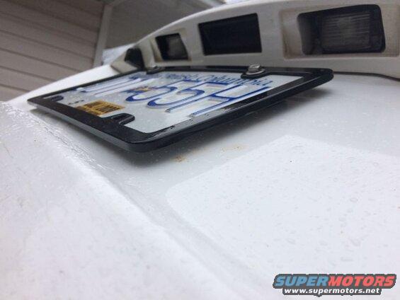 License plate frame on a white vehicle with blue lettering and a yellow sticker.