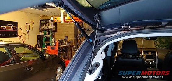 Dark blue SUV rear hatch open, showing interior and gas strut. Garage setting with another car and posters.