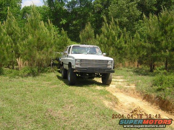 White SUV with a large grille driving on a dirt path surrounded by trees