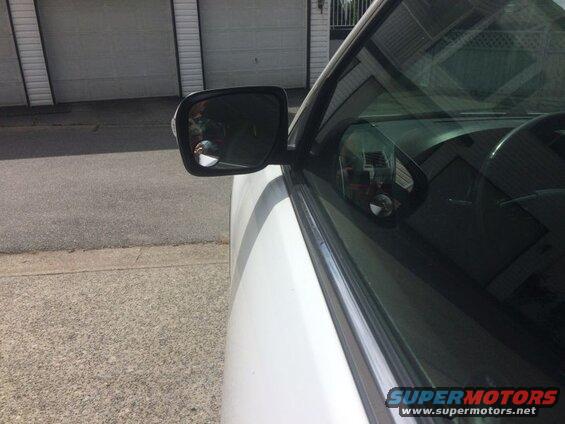 White SUV driver's side mirror reflecting a person holding a phone, interior visible through tinted window.
